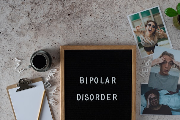 A letter board displays the words 'BIPOLAR DISORDER' surrounded by scattered letters. Nearby, there is a clipboard with a blank sheet of paper and a pencil, accompanied by a cup of black coffee. Three photographs show different facial expressions, and a small potted plant is visible. The surface is textured in a neutral tone.