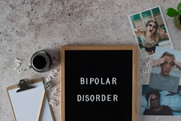 A letter board displays the words 'BIPOLAR DISORDER' surrounded by scattered letters. Nearby, there is a clipboard with a blank sheet of paper and a pencil, accompanied by a cup of black coffee. Three photographs show different facial expressions, and a small potted plant is visible. The surface is textured in a neutral tone.