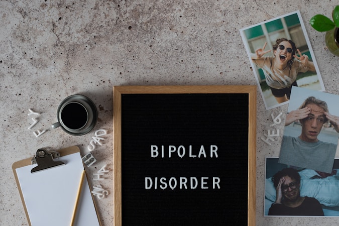 A letter board displays the words 'BIPOLAR DISORDER' surrounded by scattered letters. Nearby, there is a clipboard with a blank sheet of paper and a pencil, accompanied by a cup of black coffee. Three photographs show different facial expressions, and a small potted plant is visible. The surface is textured in a neutral tone.