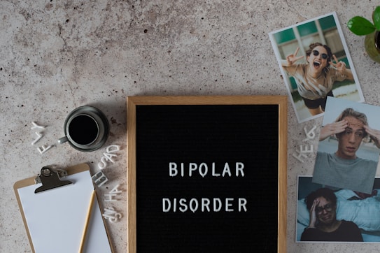 A letter board displays the words 'BIPOLAR DISORDER' surrounded by scattered letters. Nearby, there is a clipboard with a blank sheet of paper and a pencil, accompanied by a cup of black coffee. Three photographs show different facial expressions, and a small potted plant is visible. The surface is textured in a neutral tone.