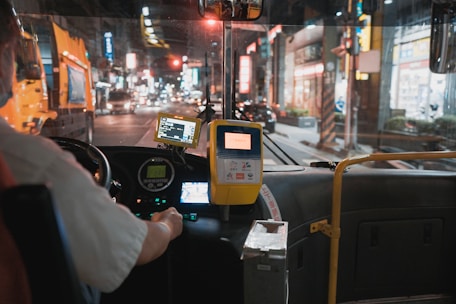 The interior view of a bus or public transportation vehicle at night from the driver's perspective. The driver is visible on the left side, steering the vehicle while a digital fare meter and several electronic devices are mounted on the dashboard. The windshield offers a view of an urban street scene with bright lights from surrounding buildings and a few vehicles on the road.