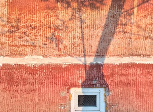 A textured wall with a reddish-orange hue is marked by horizontal lines and an uneven surface. The wall is divided by a lighter horizontal seam near the center, and the shadow of a tree branch extends across, adding depth and contrast. At the bottom, there is a small vent with a black mesh, surrounded by a light-colored rectangular frame.
