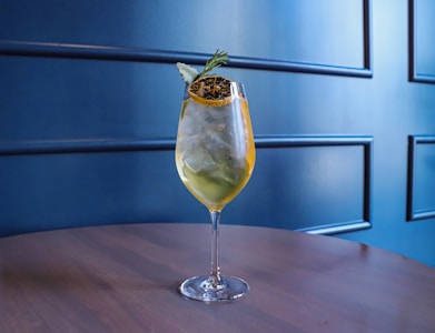A well-crafted cocktail served in a wine glass sits on a wooden table. The drink contains ice cubes and is garnished with a piece of dried citrus and a sprig of rosemary. The background features a stylish, dark blue panelled wall.
