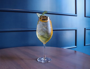 A well-crafted cocktail served in a wine glass sits on a wooden table. The drink contains ice cubes and is garnished with a piece of dried citrus and a sprig of rosemary. The background features a stylish, dark blue panelled wall.