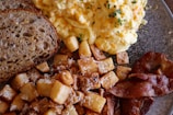 A breakfast plate featuring scrambled eggs garnished with herbs, crispy diced potatoes sprinkled with grated cheese, golden-brown bacon strips, and slices of multigrain bread.