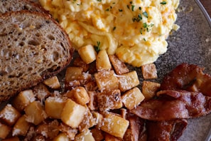 A breakfast plate featuring scrambled eggs garnished with herbs, crispy diced potatoes sprinkled with grated cheese, golden-brown bacon strips, and slices of multigrain bread.