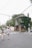 A couple walks hand in hand down a quiet street. To the right, a bride in a wedding dress is visible near a charming ivy-covered building with a cafe named 'Day & Night'. The street is lined with trees and power lines crisscross overhead.