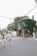 A couple walks hand in hand down a quiet street. To the right, a bride in a wedding dress is visible near a charming ivy-covered building with a cafe named 'Day & Night'. The street is lined with trees and power lines crisscross overhead.
