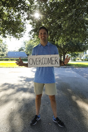A person stands outside on a paved path, holding a sign with the word 'OVERCOMER' written on it. Sunlight filters through the leaves of a large tree overhead, creating a bright and hopeful atmosphere. The person is wearing a blue shirt and beige shorts.