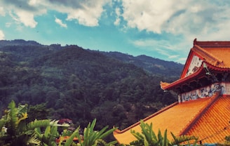 A traditional Asian temple with ornate orange roof tiles sits amidst lush green foliage with a backdrop of verdant mountains under a partly cloudy blue sky.