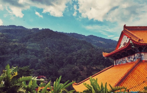 A traditional Asian temple with ornate orange roof tiles sits amidst lush green foliage with a backdrop of verdant mountains under a partly cloudy blue sky.