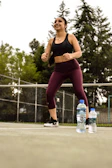 A person wearing workout attire, including a black sports bra and burgundy leggings, is exercising outdoors on a tennis court. The individual looks energetic and is smiling. In the foreground, two plastic water bottles are placed on the ground. The background features a net and tall trees, indicating an outdoor setting.