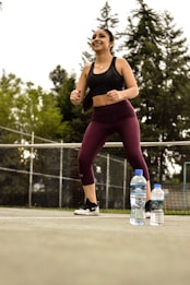A person wearing workout attire, including a black sports bra and burgundy leggings, is exercising outdoors on a tennis court. The individual looks energetic and is smiling. In the foreground, two plastic water bottles are placed on the ground. The background features a net and tall trees, indicating an outdoor setting.