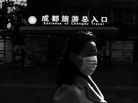 A person wearing a mask walks past a building with a sign that reads 'Entrance of Chengdu Travel' in both English and Chinese. The background features various posters and displays, while the lighting creates strong contrasts, casting parts of the scene into shadow.