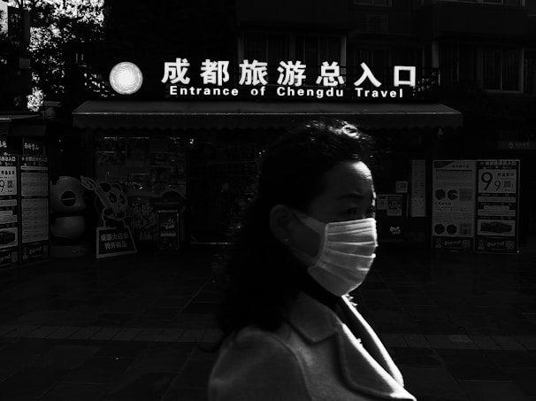 A person wearing a mask walks past a building with a sign that reads 'Entrance of Chengdu Travel' in both English and Chinese. The background features various posters and displays, while the lighting creates strong contrasts, casting parts of the scene into shadow.