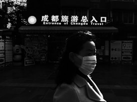 A person wearing a mask walks past a building with a sign that reads 'Entrance of Chengdu Travel' in both English and Chinese. The background features various posters and displays, while the lighting creates strong contrasts, casting parts of the scene into shadow.