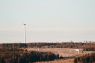 A landscape with a wind turbine standing tall against a pale sky, surrounded by a sprawling forest. Below, there is a highway with several vehicles, including a truck and cars, traveling along it.