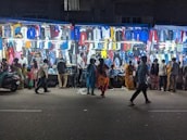 A bustling night market street scene with various clothes displayed on brightly lit stalls. Several people are walking by or browsing the stalls, examining the clothing items hanging in rows. The atmosphere is lively with a mix of vibrant colors from the clothes.