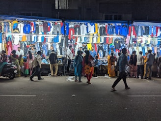 A bustling night market street scene with various clothes displayed on brightly lit stalls. Several people are walking by or browsing the stalls, examining the clothing items hanging in rows. The atmosphere is lively with a mix of vibrant colors from the clothes.