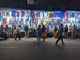 A bustling night market street scene with various clothes displayed on brightly lit stalls. Several people are walking by or browsing the stalls, examining the clothing items hanging in rows. The atmosphere is lively with a mix of vibrant colors from the clothes.