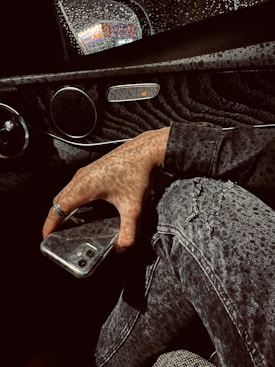 A hand holding a smartphone is visible near the console of a car. The interior features textured surfaces, and raindrops can be seen on a window. There is a seat warmer button and a view out the wet window in the background. The person's clothing consists of denim jeans and a patterned jacket.