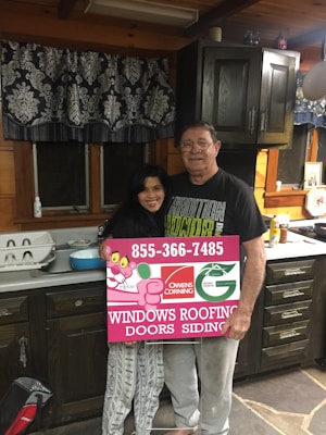 A man and woman stand in a rustic kitchen with wooden cabinets and a patterned curtain. They are holding a large rectangular sign advertising windows, roofing, doors, and siding, featuring a logo and contact number. The kitchen counter has various items, including a dish rack and some utensils.