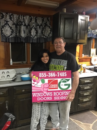 A man and woman stand in a rustic kitchen with wooden cabinets and a patterned curtain. They are holding a large rectangular sign advertising windows, roofing, doors, and siding, featuring a logo and contact number. The kitchen counter has various items, including a dish rack and some utensils.