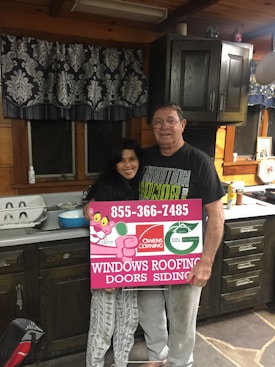 A man and woman stand in a rustic kitchen with wooden cabinets and a patterned curtain. They are holding a large rectangular sign advertising windows, roofing, doors, and siding, featuring a logo and contact number. The kitchen counter has various items, including a dish rack and some utensils.