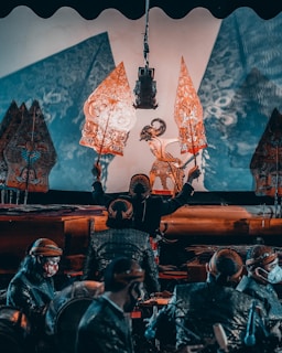 A traditional puppet performance featuring intricately designed shadow puppets held by performers in a dimly lit setting. The performers, wearing traditional attire, manipulate the puppets against a lighted backdrop, creating detailed shadows that are projected onto a screen. The scene conveys a cultural and artistic atmosphere.