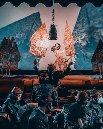 A traditional puppet performance featuring intricately designed shadow puppets held by performers in a dimly lit setting. The performers, wearing traditional attire, manipulate the puppets against a lighted backdrop, creating detailed shadows that are projected onto a screen. The scene conveys a cultural and artistic atmosphere.