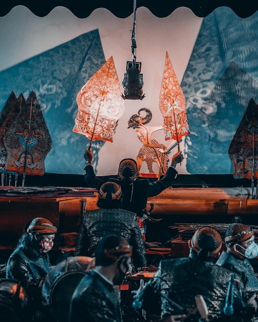 A traditional puppet performance featuring intricately designed shadow puppets held by performers in a dimly lit setting. The performers, wearing traditional attire, manipulate the puppets against a lighted backdrop, creating detailed shadows that are projected onto a screen. The scene conveys a cultural and artistic atmosphere.
