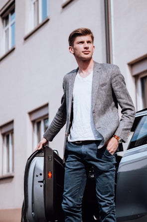 A man in a casual yet smart outfit is standing next to an open car door with his hand resting on it. He is wearing a light gray blazer, white t-shirt, and light blue pants. The background shows a building with windows, giving an urban setting.