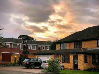 A suburban neighborhood with brick houses featuring sloped shingle roofing and large windows. There are two parked cars in a driveway, and the sky above is filled with dramatic clouds illuminated by the setting or rising sun, casting a warm glow over the scene.