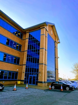A modern office building with large blue-tinted glass windows and brick walls is seen from a low angle. Several cars are parked in the adjacent parking lot, with traffic cones placed around. The sky is clear and bright, casting reflections on the glass facade.