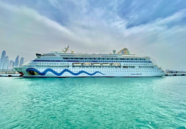 A large, white cruise ship is docked at a harbor, with a whimsical blue wave design along its side and distinctive eye motifs at the bow. Clear, turquoise water surrounds the ship, and a skyline of tall modern buildings is visible in the distance under a cloud-filled sky.