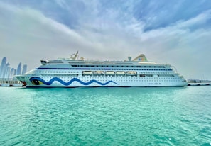 A large, white cruise ship is docked at a harbor, with a whimsical blue wave design along its side and distinctive eye motifs at the bow. Clear, turquoise water surrounds the ship, and a skyline of tall modern buildings is visible in the distance under a cloud-filled sky.