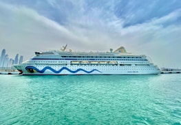A large, white cruise ship is docked at a harbor, with a whimsical blue wave design along its side and distinctive eye motifs at the bow. Clear, turquoise water surrounds the ship, and a skyline of tall modern buildings is visible in the distance under a cloud-filled sky.