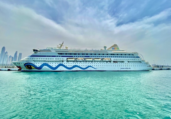 A large, white cruise ship is docked at a harbor, with a whimsical blue wave design along its side and distinctive eye motifs at the bow. Clear, turquoise water surrounds the ship, and a skyline of tall modern buildings is visible in the distance under a cloud-filled sky.