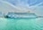 A large, white cruise ship is docked at a harbor, with a whimsical blue wave design along its side and distinctive eye motifs at the bow. Clear, turquoise water surrounds the ship, and a skyline of tall modern buildings is visible in the distance under a cloud-filled sky.