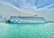 A large, white cruise ship is docked at a harbor, with a whimsical blue wave design along its side and distinctive eye motifs at the bow. Clear, turquoise water surrounds the ship, and a skyline of tall modern buildings is visible in the distance under a cloud-filled sky.