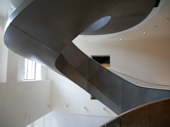 A modern architectural interior featuring a large, sweeping staircase with smooth curves and a metallic finish. The surrounding space has a minimalistic design with neutral-colored walls and ceiling lights providing subtle illumination. Large windows allow natural light to enter, enhancing the clean, sleek aesthetic.