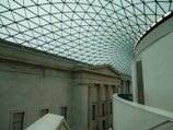 An interior view of a large building with a classical architectural design. The space features a high, curved glass ceiling with a geometric pattern. Below, the walls are made of stone with columns and extensive molding. The lighting is soft and ambient, enhancing the spaciousness of the area.