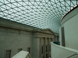 An interior view of a large building with a classical architectural design. The space features a high, curved glass ceiling with a geometric pattern. Below, the walls are made of stone with columns and extensive molding. The lighting is soft and ambient, enhancing the spaciousness of the area.