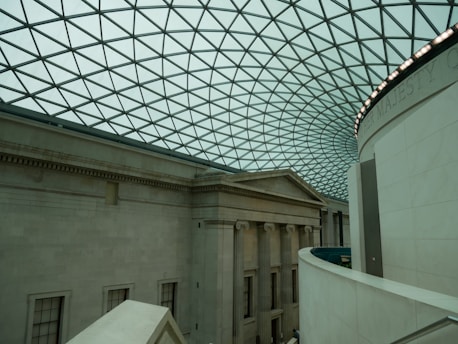An interior view of a large building with a classical architectural design. The space features a high, curved glass ceiling with a geometric pattern. Below, the walls are made of stone with columns and extensive molding. The lighting is soft and ambient, enhancing the spaciousness of the area.