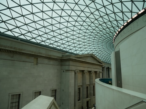 An interior view of a large building with a classical architectural design. The space features a high, curved glass ceiling with a geometric pattern. Below, the walls are made of stone with columns and extensive molding. The lighting is soft and ambient, enhancing the spaciousness of the area.
