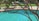 A backyard swimming pool with clear blue water, partially shaded by the branches of a tree in the foreground. Patio furniture including cushioned chairs is visible near the pool area, situated on a concrete deck surrounded by green grass.