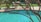 A backyard swimming pool with clear blue water, partially shaded by the branches of a tree in the foreground. Patio furniture including cushioned chairs is visible near the pool area, situated on a concrete deck surrounded by green grass.
