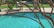 A backyard swimming pool with clear blue water, partially shaded by the branches of a tree in the foreground. Patio furniture including cushioned chairs is visible near the pool area, situated on a concrete deck surrounded by green grass.