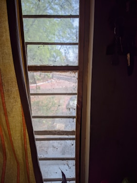 A barred window with a mesh screen overlooks a view of a tree and yard. The curtain on the left has vertical stripes in earthy tones, and there are some keys hanging on the right. The outside view is slightly obscured by the grime and dirt on the screen.