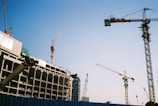 Multiple construction cranes are visible at a building site. A partially constructed building appears to the left, with cranes extended into the sky. The blue sky provides a clear backdrop, emphasizing the scale and complexity of the construction process.